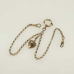 Puede incluir: Una cadena de reloj dorada con un dije en forma de corazón. La cadena presenta una serie de pequeños círculos enlazados e incluye un cierre en cada extremo. El dije de corazón tiene una superficie texturizada y un pequeño acento transparente.