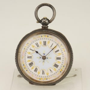 Puede incluir: Reloj de bolsillo antiguo con caja plateada y esfera blanca con números romanos negros y detalles dorados. El reloj tiene un bisel decorativo y un asa para una cadena.