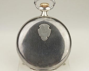 Peut inclure: Une montre de poche argentée avec un motif à rayures verticales au dos. La montre a une boucle argentée et une couronne dorée. La montre est exposée sur un support transparent.