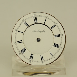 Puede incluir: Una esfera de reloj vintage con fondo blanco, números romanos negros y una pista de minutos. Las palabras "Non-Magnetic" están impresas en negro. La esfera del reloj está colocada sobre un soporte transparente y rectangular.