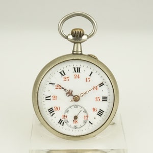 Puede incluir: Un reloj de bolsillo plateado con esfera blanca, números romanos negros y números arábigos rojos. El reloj tiene un pequeño dial de segundos y manecillas ornamentadas. La corona es dorada. El reloj se exhibe sobre un soporte transparente.