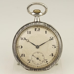 Puede incluir: Un reloj de bolsillo plateado con esfera blanca y agujas negras. El reloj tiene un pequeño dial de segundos y una esfera texturizada. El reloj tiene un lazo plateado y una corona dorada.