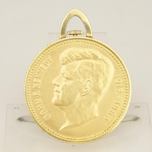 Puede incluir: Colgante dorado con un perfil detallado de John F. Kennedy. El diseño, similar a una moneda, incluye la inscripción "JOHN F. KENNEDY" y los años "1917-1963". El colgante tiene un borde decorativo y un pequeño lazo en la parte superior para una cadena.