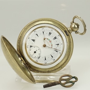 Puede incluir: Un reloj de bolsillo antiguo con esfera blanca, manecillas negras y un pequeño dial de segundos. El reloj tiene una caja dorada y una llave. La esfera tiene el texto "DENT LONDON". La tapa del reloj está abierta.