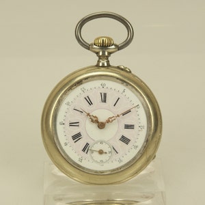 Puede incluir: Un reloj de bolsillo plateado con esfera blanca, números romanos negros y manecillas doradas. El reloj tiene un pequeño dial de segundos y un lazo plateado. La esfera tiene un diseño floral rosa.