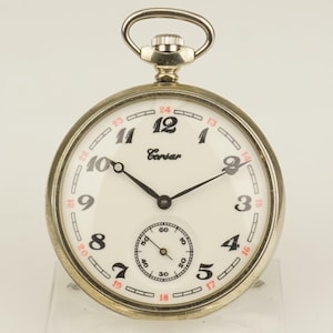 Puede incluir: Un reloj de bolsillo plateado con esfera blanca. Los números negros marcan las horas, con números rojos para los minutos exteriores. La esfera del reloj tiene un dial de segundos más pequeño. La marca "Corsar" está impresa en la esfera.