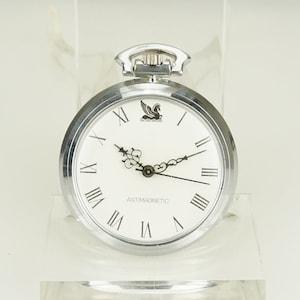 Puede incluir: Un reloj de bolsillo plateado con esfera blanca y números romanos negros. El reloj presenta un emblema negro encima del centro y la palabra "ANTIMAGNETIC" debajo. El reloj se exhibe sobre un soporte transparente.