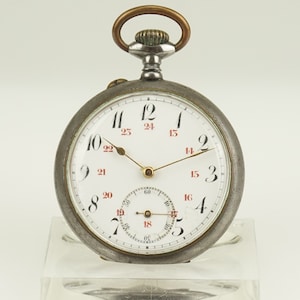 Puede incluir: Un reloj de bolsillo vintage con esfera blanca y números negros y rojos. El reloj tiene un pequeño dial de segundos y una caja de metal con corona y asa. Las manecillas son doradas.