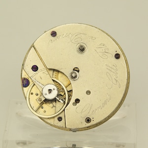 Puede incluir: Primer plano de un movimiento de reloj antiguo con intrincados engranajes y componentes. El metal plateado está inscrito con una elegante escritura cursiva, incluido el nombre "Gille Clement". Un ejemplo de artesanía de precisión.
