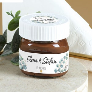 Könnte beinhalten: Ein kleines Glas gefüllt mit Schokoladenaufstrich, mit einem weißen Deckel und einem personalisierten Etikett. Das Etikett zeigt ein florales Design mit grünen Blättern und dem Text "Elena & Stefan 16.09.2023".