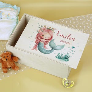 Peut inclure: Coffret en bois clair avec couvercle à charnière. Le couvercle présente une illustration de sirène de dessin animé avec des cheveux roses et une queue turquoise, ainsi que le nom "Emilia" et la date "28.11.2024". Un petit ours en peluche et une sucette sont visibles.