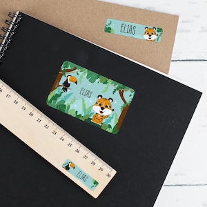 Può includere: Un set di tre etichette per il nome personalizzate con un tema giungla. Le etichette presentano un tucano, una tigre e il nome "ELIAS" in uno schema di colori blu e verde.