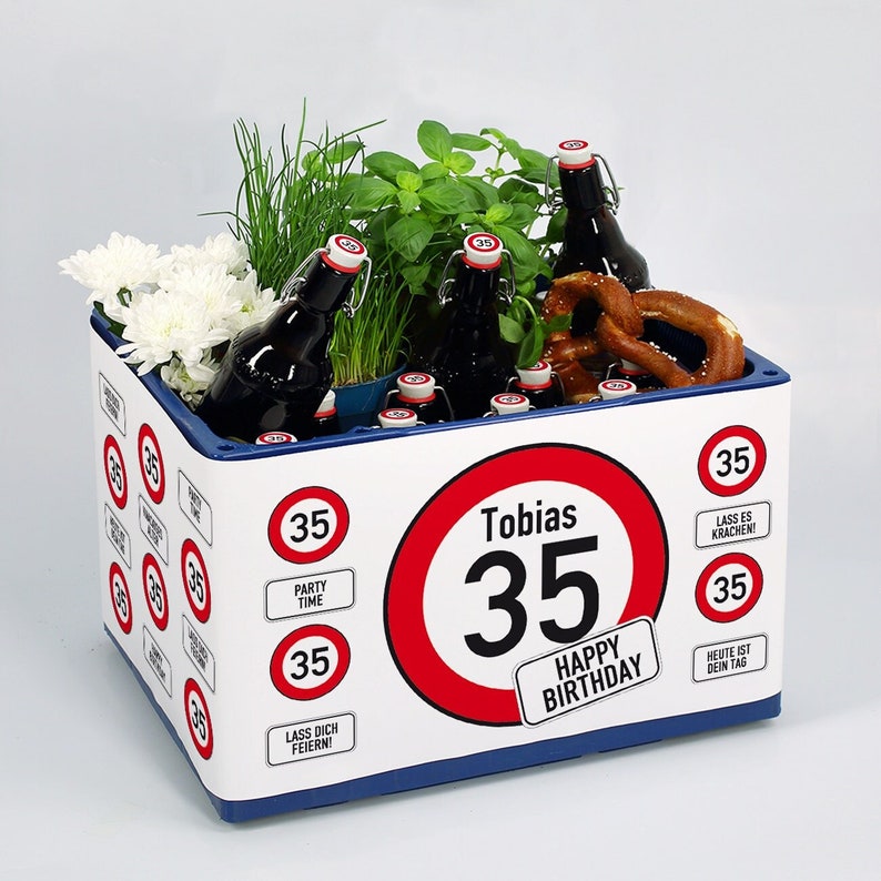 May include: A white gift box with a blue lid decorated with red and white speed limit signs. The signs feature the number 35 and various phrases like "Happy Birthday", "Party Time", and "Lass es krachen!" The box is filled with beer bottles, a pretzel, and greenery.