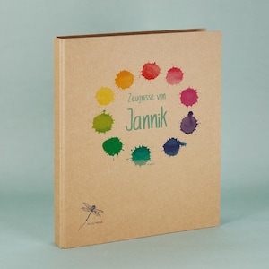 Może przedstawiać: Notatnik w twardej oprawie z okładką z papieru kraft. Okładka zawiera kolorowy wzór rozprysków akwareli wokół imienia "Jannik" w kolorze zielonym. Tekst "Zeugnisse von" znajduje się nad imieniem. Ilustracja ważki znajduje się w lewym dolnym rogu.