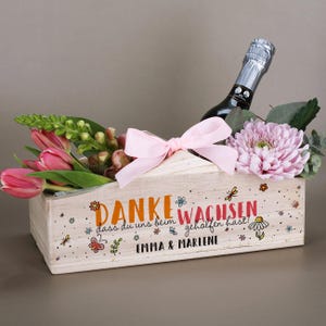 Può includere: Una fioriera rettangolare in legno con un fiocco rosa, riempita di tulipani rosa, un crisantemo viola chiaro e verde. Una bottiglia di spumante è inserita all'interno. La scatola riporta il testo "DANKE DASS DU UNS BEIM WACHSEN GEHOLFEN HAST!" e "EMMA & MARLENE" con illustrazioni floreali colorate.