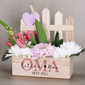 Könnte beinhalten: Eine Holzkiste mit Zaun-Design, gefüllt mit rosa Tulpen, weißen und rosa Chrysanthemen und Grünzeug. Ein hellvioletter herzförmiger Anhänger ist am Zaun befestigt. Auf der Kiste stehen die Worte "OMA", die Namen "Celina Neo" und das Jahr "2011".