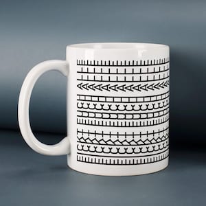 Könnte beinhalten: Ein weißer Keramikbecher mit einem sich wiederholenden schwarzen Grafikmuster aus horizontalen Bändern. Das Design besteht aus verschiedenen geometrischen Formen, darunter vertikale Linien, Dreiecke und geschwungene Motive, die in Reihen angeordnet sind.