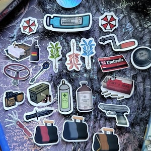 May include: A collection of stickers featuring various designs. Includes stickers of a T-Virus vial, Umbrella Corporation logos, a key, a gun, and other themed graphics. The stickers are colorful and detailed, with some featuring text.