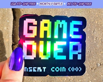 May include: A black and iridescent sticker with the words "GAME OVER" in pixelated rainbow letters. The sticker also has the text "INSERT COIN (coo)" in white letters.