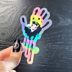 May include: A holographic sticker of a black handprint with dripping rainbow paint. The sticker is on a dark wood background.
