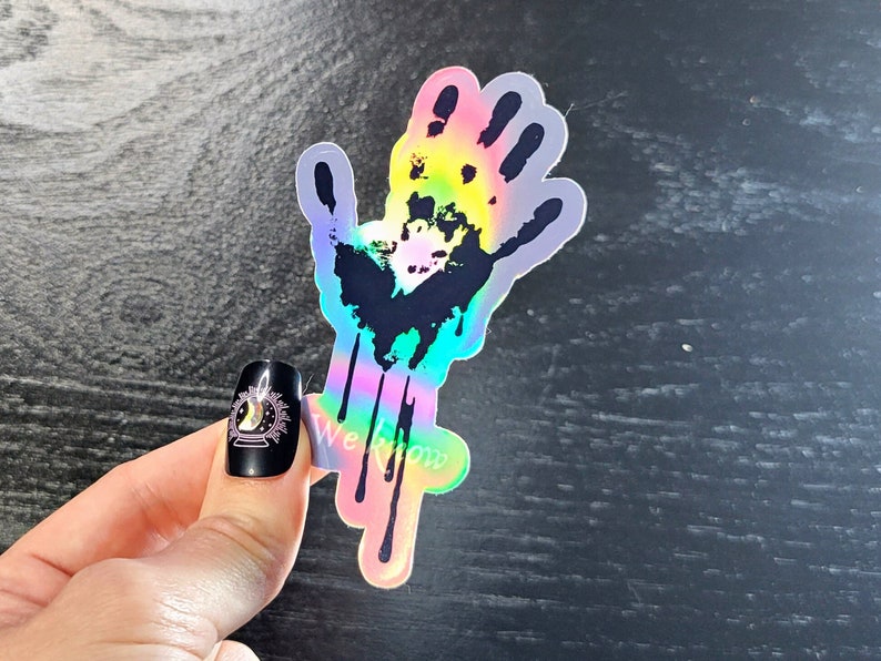 May include: A holographic sticker of a black handprint with dripping rainbow paint. The sticker is on a dark wood background.