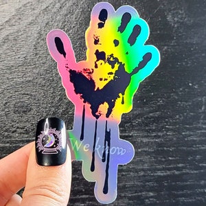 May include: A holographic sticker of a black handprint with dripping paint. The handprint is outlined in rainbow colors. The sticker has the text "We Know" at the bottom.