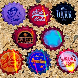 May include: A collection of eight bottle caps with various designs and text. The caps feature the words "Nuka Cola," "Sunset Sarsaparilla," "Nuka Dark," "Nuka Orange," "Nuka Quantum," and "Nuka Cherry." The caps have a metallic, iridescent finish.
