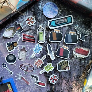 May include: A collection of stickers featuring various designs, including a red and white umbrella logo, a vial labeled "T-VIRUS", and a handgun. Other stickers depict a camera, a spray can, and a toolbox. The stickers are arranged on a dark, patterned surface.