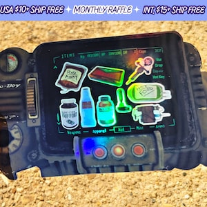 May include: A Pip-Boy 3000 replica with a screen displaying a Fallout video game inventory. The screen shows various items, including Rad-Away, Stimpak, and a bottle of Nuka-Cola. The Pip-Boy is made of black plastic and has a silver metal trim.