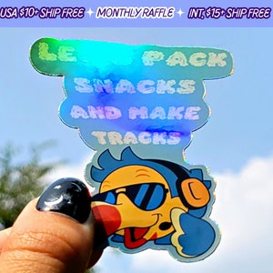 May include: A holographic sticker with a cartoon bird wearing sunglasses and headphones. The sticker says "LE PACK SNACKS AND MAKE TRACKS".