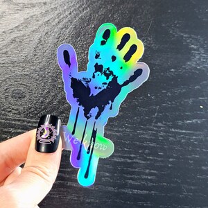 May include: A holographic sticker of a black handprint with dripping paint. The sticker has a rainbow iridescent finish.