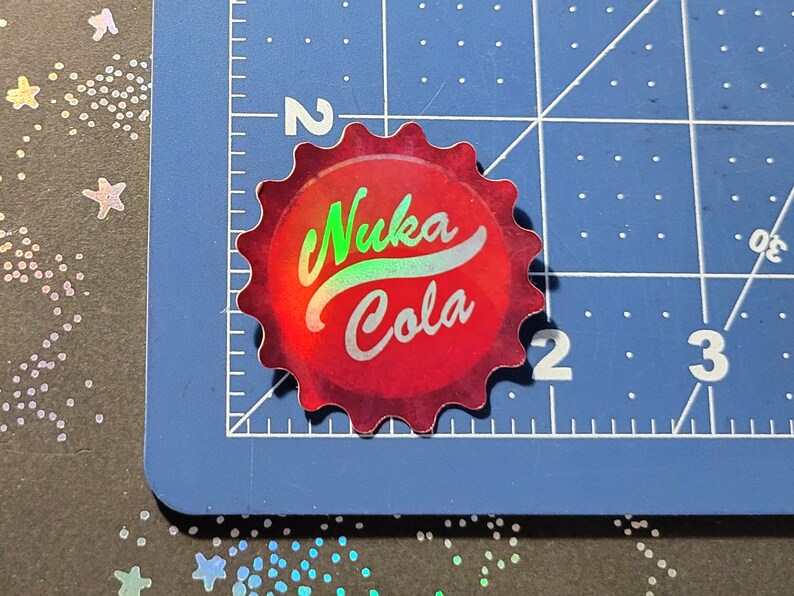 May include: A red, bottle cap-shaped sticker with the words "Nuka Cola" in a holographic, iridescent font. The sticker is on a blue cutting mat with a ruler and numbers.