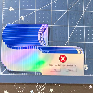 May include: A holographic sticker with a blue and white color scheme. The sticker is a parody of a Windows error message. The text on the sticker reads "Error: Task failed successfully. Ok Cancel".