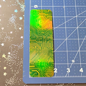 May include: A rectangular bookmark with a circuit board design. The bookmark features a green and yellow color scheme with intricate lines and patterns, resembling a computer circuit. The top edge has a gradient of green to orange.