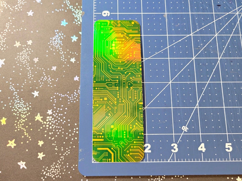 May include: A rectangular bookmark with a circuit board design. The bookmark features a green and yellow color scheme with intricate lines and patterns, resembling a computer circuit. The top edge has a gradient of green to orange.