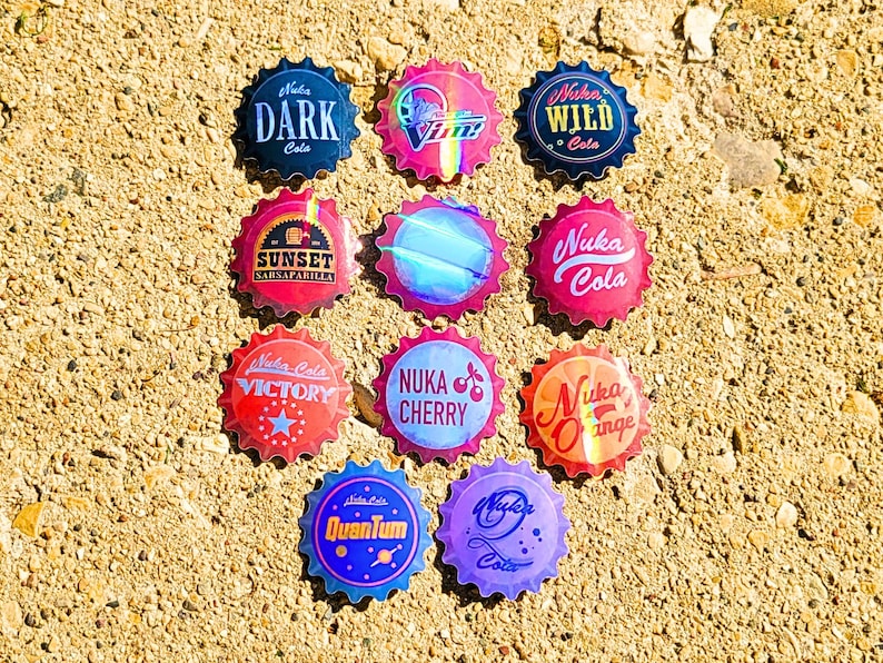 May include: A collection of colorful bottle caps with various designs and text, including "Nuka Cola," "Dark Cola," and "Sunset Sarsaparilla." The caps are arranged on a textured, light brown surface.