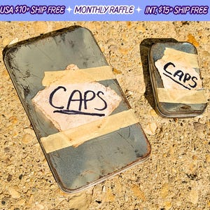 May include: Two vintage metal tins with the word "CAPS" written on them in black marker. The tins are sitting on a sandy surface.