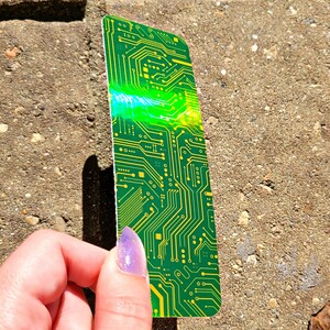 May include: A green and yellow circuit board design bookmark. The bookmark has a rectangular shape with rounded corners. The design features intricate lines and patterns, with a bright green and yellow color scheme. The bookmark is held by a hand.