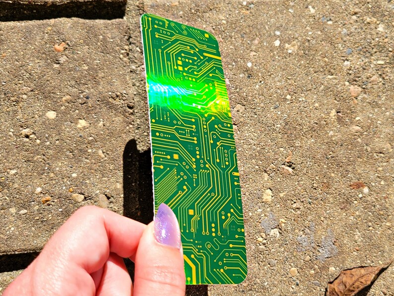 May include: A green and yellow circuit board design bookmark. The bookmark has a rectangular shape with rounded corners. The design features intricate lines and patterns, with a bright green and yellow color scheme. The bookmark is held by a hand.