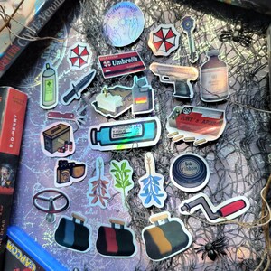 May include: A collection of stickers with various designs, including the Umbrella Corporation logo, a handgun, a vial labeled "T-Virus", and other themed graphics. The stickers are arranged on a purple map background.