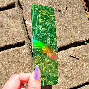 May include: A green and yellow circuit board design bookmark. The bookmark has a rectangular shape with rounded corners. The design features intricate yellow lines and shapes on a green background. The bookmark is held by a hand with purple nail polish.