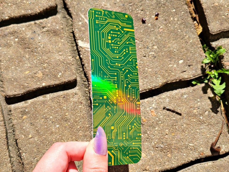 May include: A green and yellow circuit board design bookmark. The bookmark has a rectangular shape with rounded corners. The design features intricate yellow lines and shapes on a green background. The bookmark is held by a hand with purple nail polish.