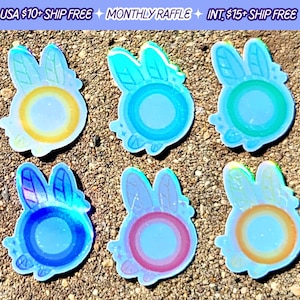 May include: Six iridescent holographic stickers featuring a cartoon bunny with a halo and a circle in the center. The stickers are on a brown sandy surface.