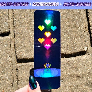 May include: A holographic bookmark featuring pixelated hearts in various colors, with a flower at the bottom. The text "MONTHLY RAFFLE" is visible at the top. The bookmark has a dark blue background with a rainbow effect.