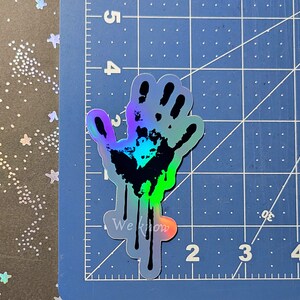 May include: A holographic sticker of a handprint with dripping paint. The handprint is black and the paint is a gradient of blue, green, and purple. The sticker has the text "We Know" printed on it.