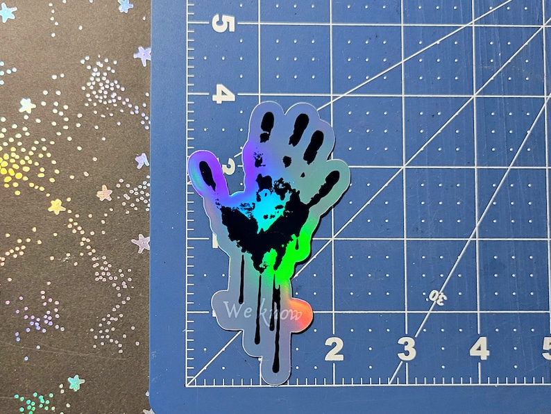 May include: A holographic sticker of a handprint with dripping paint. The handprint is black and the paint is a gradient of blue, green, and purple. The sticker has the text "We Know" printed on it.