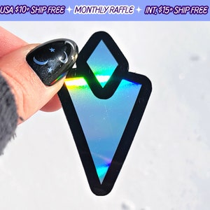 May include: A holographic sticker with a black outline in the shape of a diamond. The sticker is iridescent and reflects light in a rainbow of colors.