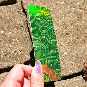 May include: A rectangular bookmark with a circuit board design. The bookmark features a green and yellow color scheme with intricate lines and patterns, resembling the internal components of a computer. The edges have a gradient of green to orange.