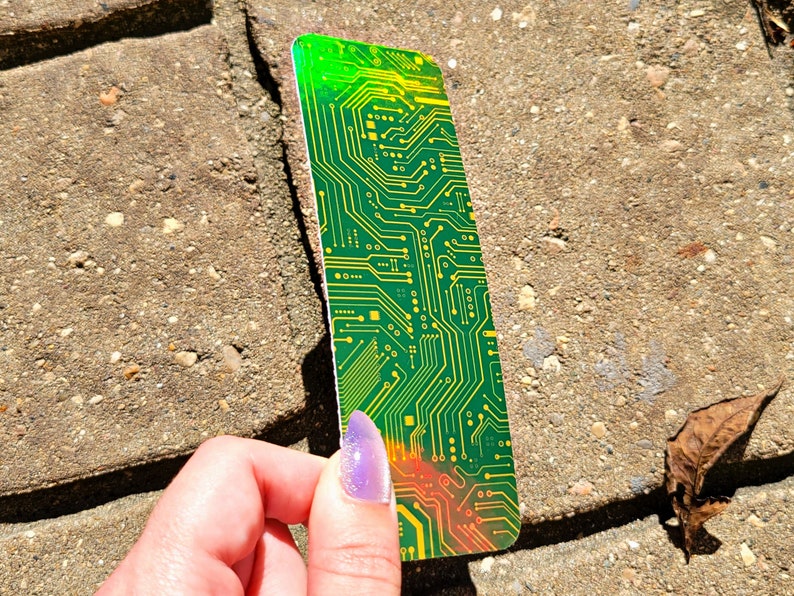 May include: A rectangular bookmark with a circuit board design. The bookmark features a green and yellow color scheme with intricate lines and patterns, resembling the internal components of a computer. The edges have a gradient of green to orange.