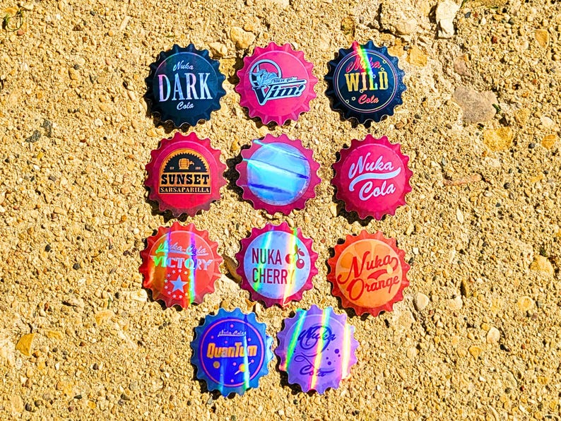 May include: A collection of colorful bottle caps with various designs and text, including "DARK Cola," "Sunset Sarsaparilla," and "Nuka Cola." The caps are arranged on a textured, sandy surface.